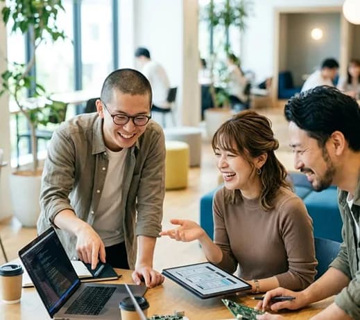 Three colleagues laughing together over a laptop and tablet in a bright office