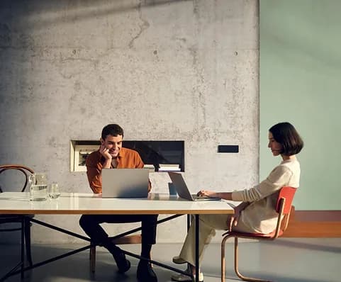 Two colleagues working on laptops at a long table in a concrete-walled studio