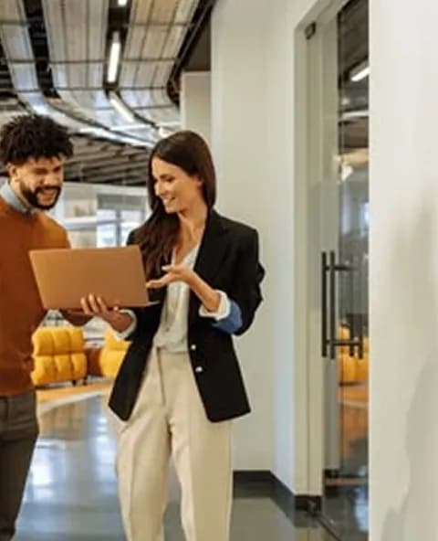 Two colleagues smiling over a laptop in an office corridor