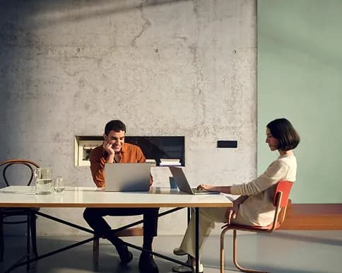 Two colleagues working on laptops at a long table in a concrete-walled room
