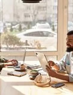 A man reading by a sunny café window beside a laptop