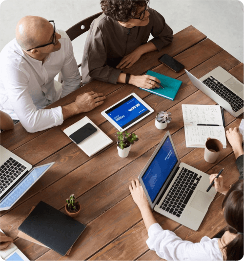 Top-down view of a team collaborating around a wooden meeting table with laptops, tablets, and notes