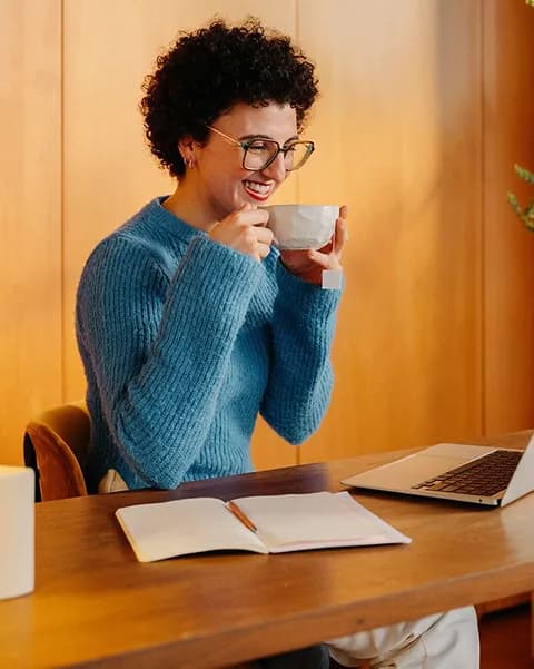 A smiling woman sipping coffee at a desk with a notebook and laptop