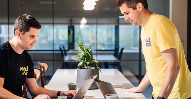 Two colleagues smiling while working on laptops at a long table